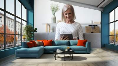 Smiling middle aged woman sitting on comfortable sofa with computer digital tablet. Happy older generation lady shopping online in internet store at home, using electronic device for communication. Wall mural