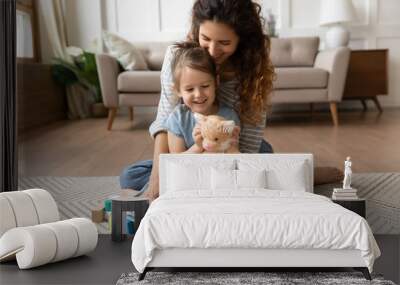 Smiling mother and little daughter hugging, playing with toys, sitting on warm floor with underfloor heating in living room, happy young mum and preschool girl enjoying leisure time together Wall mural