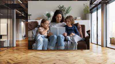 Smiling mother reading book to adorable little kids sitting on cozy couch at home, loving mom with little son and daughter engaged in educational activity, family spending leisure time together Wall mural