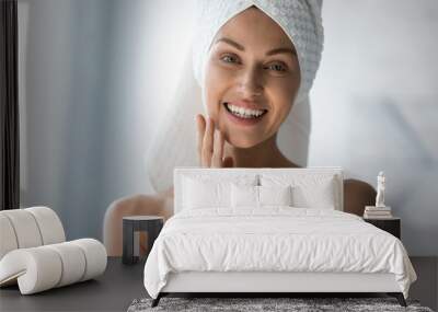 Smiling pretty young woman with towel on head looking at camera, touching cheek. Happy beautiful lady satisfied with moisturized clean skin condition, enjoying morning skincare routine in bathroom. Wall mural