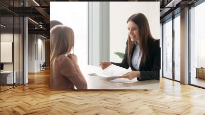 Smiling real estate agent offering to young couple to buy a cottage while meeting at office, presenting technical documentation such as building house plan, telling about advantages of country living Wall mural