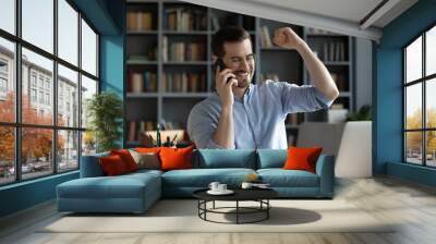 Smiling satisfied businessman wearing glasses making phone call, hearing good news, job promotion celebrating success, showing yes gesture, sitting at work desk with laptop and using smartphone Wall mural