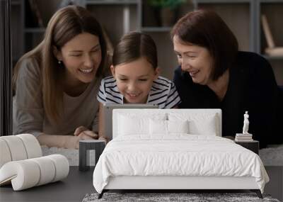 Smiling three generations of Caucasian women relax at home together watch funny video on tablet. Happy little teen girl child with young mother and old grandmother use modern pad device playing. Wall mural