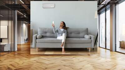 Smiling woman using air conditioner remote controller, sitting on cozy couch at home full length, happy young female relaxing setting comfort temperature in modern living room, enjoy fresh air Wall mural