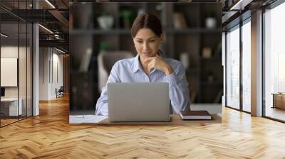 Smiling young Caucasian businesswoman sit at desk at home office look at laptop screen consult client online. Millennial female employee or woman worker work distant on computer, analyze paperwork. Wall mural