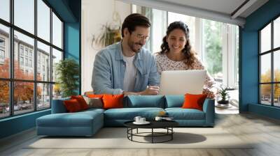 Smiling young Caucasian couple sit at desk at home look at laptop screen shopping online together. Happy husband and wife browse wireless internet on computer. Technology, communication concept. Wall mural