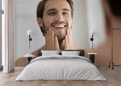 Smiling young Caucasian man look in mirror in bathroom touch healthy face skin after treatment or procedure, happy millennial male satisfied with beard or bristle after shaving, body hygiene concept Wall mural