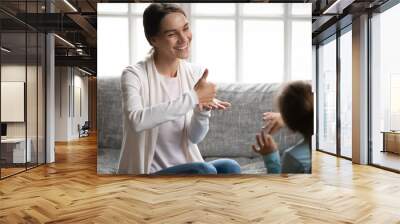 Smiling young Caucasian mom or nanny learn sign language with little disabled girl child at home. Happy positive teacher or tutor make hand gesture talk communicate nonverbal with small kid. Wall mural