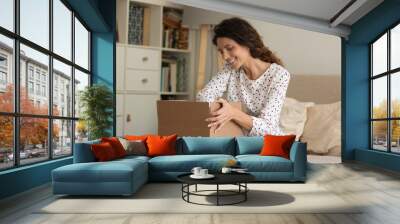 Smiling young Caucasian woman sit on sofa at home unbox package with internet order. Happy excited millennial female client or buyer unpack box shopping online. Good delivery service concept. Wall mural