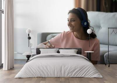 Smiling young ethnic woman in wireless headphones study online on training or course use laptop make notes, happy indian Arabic female in earphones look at computer screen watch webinar on gadget Wall mural