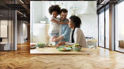 Smiling young european man holding on hands pretty small biracial adopted girl daughter, watching happy african american mother preparing healthy food, chopping fresh vegetables for salad in kitchen. Wall mural