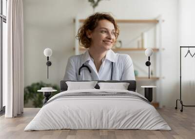 Smiling young female professional doctor holding digital tablet looking away sitting at workplace desk. Happy beautiful woman physician wearing white coat and stethoscope dreaming in medical office. Wall mural