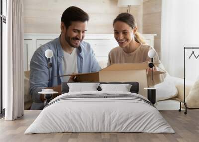 Smiling young man and woman have fun open unpack box shopping online together on internet. Happy multiracial couple buyers feel excited with good delivery service, unbox package with order. Wall mural