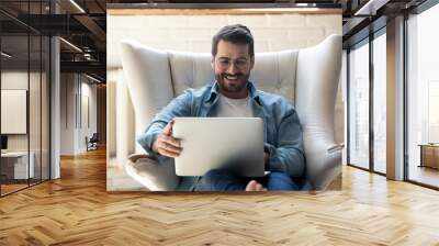 Smiling young man wearing glasses using laptop, sitting in cozy armchair, happy male looking at computer screen, chatting in social network or shopping online, playing game, working at home Wall mural