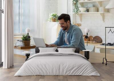 Smiling young man working on laptop in modern kitchen, checking email in morning, writing message in social network, happy young male using internet banking service, searching information Wall mural