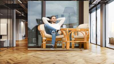 Smiling young relaxed man enjoying pleasant morning sitting on rattan chair on terrace outdoor, happy successful businessman rests outside luxury house in peaceful place, stress free weekend concept Wall mural