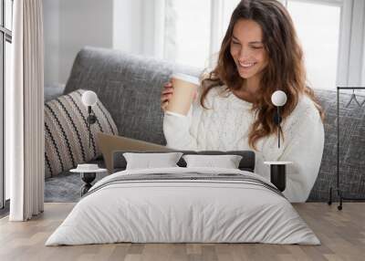 Smiling young woman enjoying morning coffee using laptop sitting at home on sofa, attractive happy girl  typing on computer having fun online or chatting with friends while relaxing on couch Wall mural