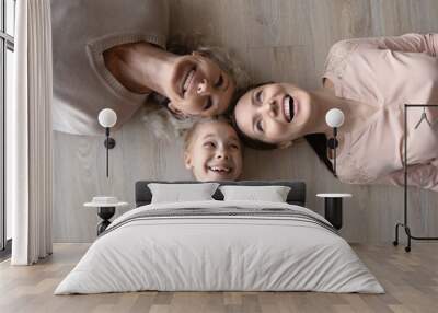 Star of 3 rays. Funny top view family portrait of three diverse women kid daughter young mother senior grandma lying head to head. Laughing three female generations having fun posing on heating floor Wall mural