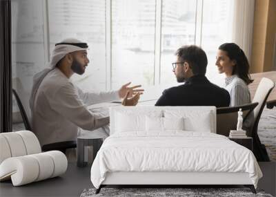 Successful young Arabian business owner man in white Muslim clothes talking to European partners at office meeting table, discussing collaboration, partnership terms, planning teamwork Wall mural