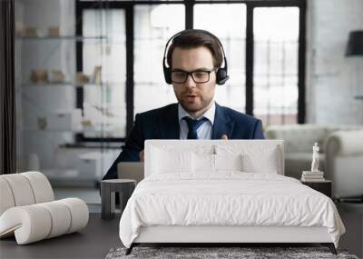 Successful young Caucasian businessman in earphones look at laptop screen consult business client or partner online. Male CEO or boss in headphones work online on computer talk on video call. Wall mural