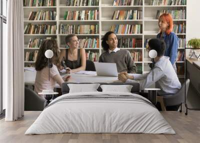 Teen red haired student girl standing at library table, speaking to diverse group of friends studying in college campus, working on class project, sitting together, talking, laughing Wall mural