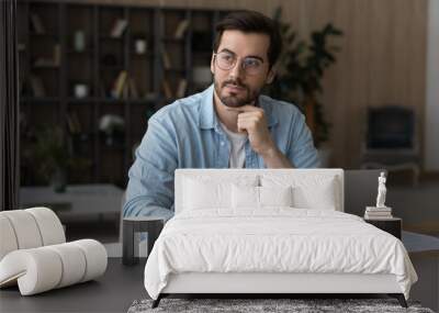 Thoughtful dreamy businessman in glasses sitting at work desk with laptop, looking to aside, pensive young man freelancer or student pondering project strategy, planning, visualizing future Wall mural