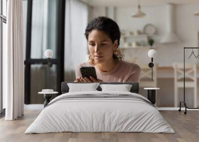 Thoughtful latin woman look at cell screen ponder on web store sales proposition reflect over answer on business offer. Interested young lady mobile phone user sit on sofa download new app for testing Wall mural