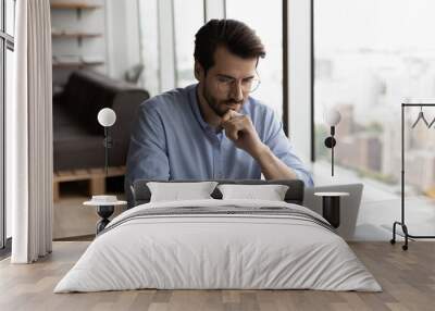 Thoughtful serious male freelancer sit at table work on computer look at screen pondering or making decision. Pensive man employee worker busy at laptop, brainstorm think over problem solution. Wall mural