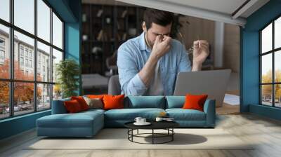 Tired young Caucasian man work on laptop at home office take off glasses overwhelmed with computer job. Exhausted male employee suffer from headache or dizziness from screen. Eyesight problem concept. Wall mural