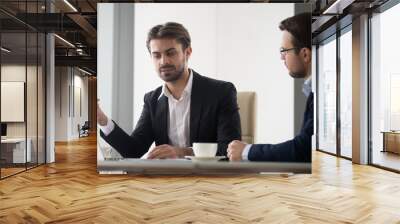 Two caucasian men in suits are discussing and planning project. Coworkers or manager and subordinate are sitting at the table and looking at monitor. One men explain arguments in favor of his opinion Wall mural