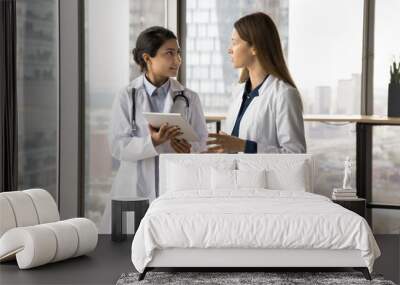 Two diverse doctor women using tablet, discussing medical case, electronic records, online healthcare service, holding digital gadget, talking, consulting each other Wall mural