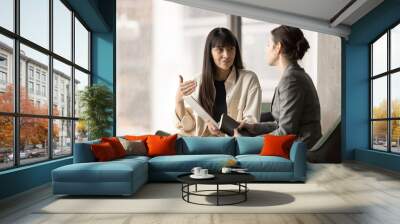 Two female office friends chatting in office lobby on work break, discussing online project, sharing tablet, speaking. Young professional listening to advice of elder business colleague Wall mural