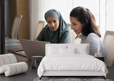 Two focused young Arab and Hispanic managers women working at laptop together, watching online work content, discussing Internet project, software product, application Wall mural