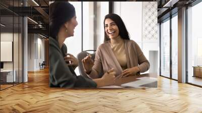 Two happy Hispanic female office friends laughing at workplace, having fun, talking at laptop, discussing funny creative ideas, brainstorming, chatting, enjoying business friendship Wall mural