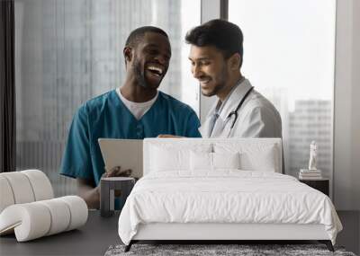 Two millennial male doctors of diverse ethnicities specialities stand by window at clinic office look on pad pc screen laugh watch funny video. Candid young medics friends having fun talk discuss work Wall mural