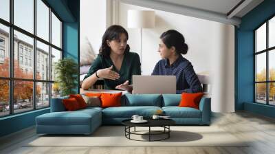 Two multi-ethnic businesswomen colleagues sit at desk, discuss joint project sit in office at workplace desk with laptop, share vision, ideas, planning cooperation. Teamwork, apprenticeship, mentoring Wall mural