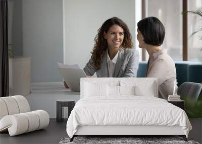 Two positive confident diverse business colleagues women talking at workplace, discussing project documents, at laptop, speaking, smiling, laughing. Mature manager consulting expert Wall mural