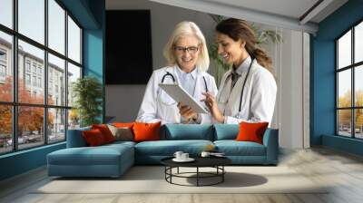 Two positive young and senior doctor women using tablet together, discussing medical application, meeting in boardroom, talking, smiling. Younger practitioner consulting elder colleague Wall mural