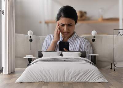 Unhappy Indian millennial woman looking at phone screen, sitting on couch, reading bad news in message, upset young female holding smartphone, having problem with broken or discharged mobile device Wall mural