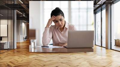 Unhappy young Indian woman sit at desk in office look at laptop screen have gadget operational problems. Upset stressed ethnic female employee work on computer frustrated with virus or scam. Wall mural