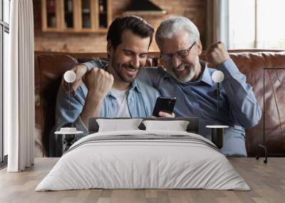 We win. Overjoyed young man and aged father laugh raise fists celebrate victory at web lottery win game on phone. Excited male pensioner and grown son watch online soccer match support football team Wall mural