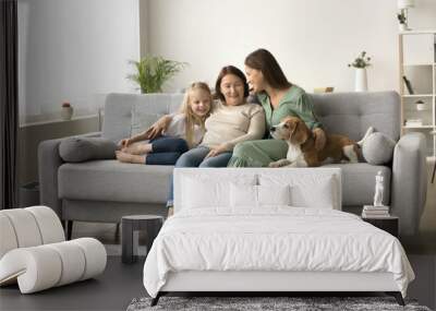 Wide family home portrait of three female generations with dog. Happy grandma, mother and little kid girl stroking cute beagle, sitting on sofa close together, talking, laughing Wall mural