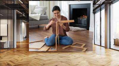 Woman assembling new furniture at home, new purchase, hobby concept. Young Indian female sit on warm floor in living room with fireplace holding wooden boards chair details, collects modern furniture Wall mural