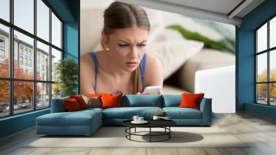 Worried young lady lying on sofa in front of laptop and looking on cellphone in her hands with confusion. Stressed lady reading unexpected message with bad news, received sad e-mail on mobile phone Wall mural