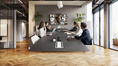 Young African American business leader holding video conference, talking to team online and offline. Workgroup sitting at meeting table, looking at large screen with headshots Wall mural