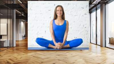 Young attractive woman practicing yoga, sitting in Butterfly exercise, baddha konasana pose, working out, wearing sportswear, blue suit, indoor full length, white loft studio background Wall mural