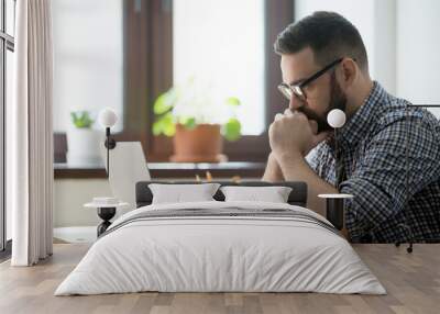 Young bearded manager working and reading data on laptop in home office. Thoughtful casual businessman thinking about job and looking on screen of notebook Wall mural