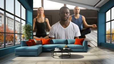 Young black man and a group of sporty people practicing yoga lesson with instructor, sitting in Sukhasana exercise, Easy Seat pose with mudra gesture, students training in club, working out, close up Wall mural