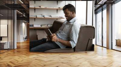 Young black man sit on sofa at modern office wait for job interview meeting appointment spend time for reading electronic book on tablet pc. Relaxed african guy hipster chat online using digital pad Wall mural