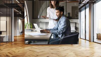 Young business assistant woman standing at Indian project manager man typing on laptop, giving professional help, talking to boss. Two colleagues working together at table with computer. Vertical shot Wall mural